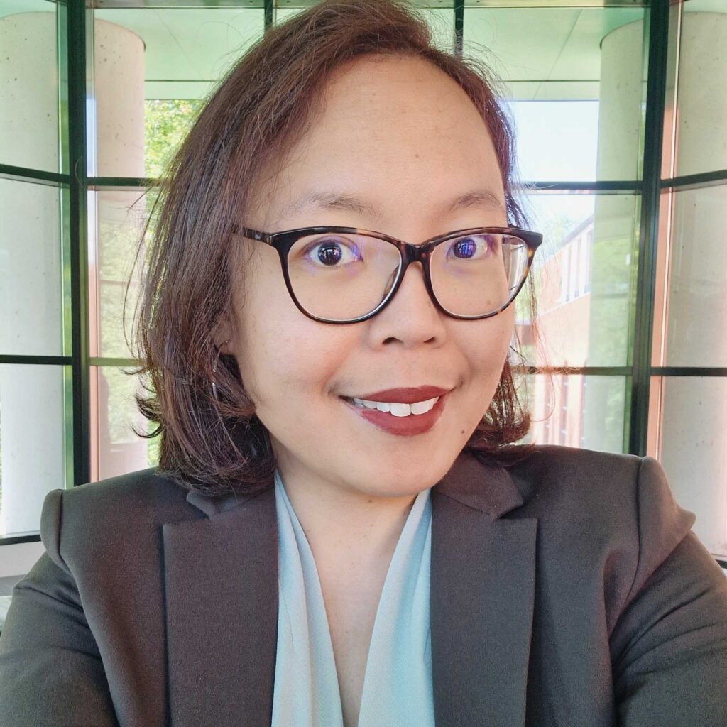 Photo of Ayu Adiati, an Indonesian woman with shoulder length dark hair, wearing dark rimmed glasses and smiling to the camera.