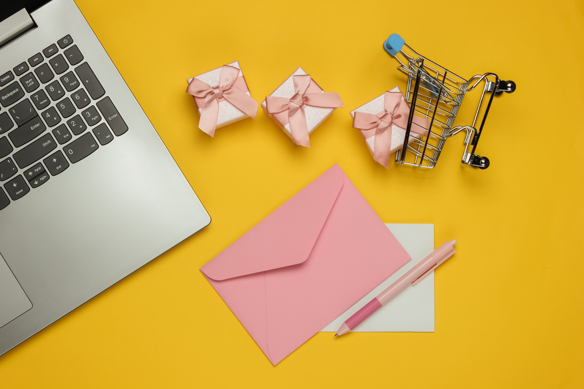 Photo of a laptop against a yellow background with the screen open, a pink envelope on the table and a wire shopping cart sideways on, spilling gifts onto the table.