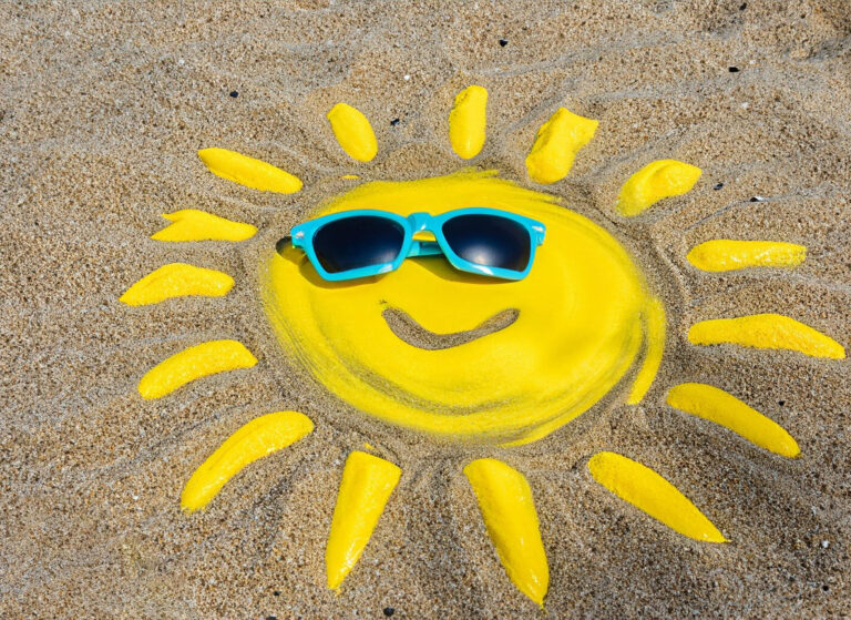 A photo of a sandy beach with a sun drawn in bright yellow sand, with a smile and a pair of blue rimmed sunglasses on it.