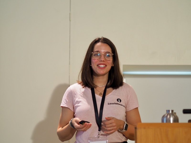 A photo of a woman standing on a stage with a remote presenter in her hand. She has a pale pink shirt on and shoulder length dark hair.